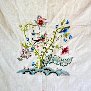 May include: A white linen tablecloth with colorful embroidered flowers and birds. The embroidery features a green vine with pink, red, and blue flowers, and two birds with blue, red, and green feathers.