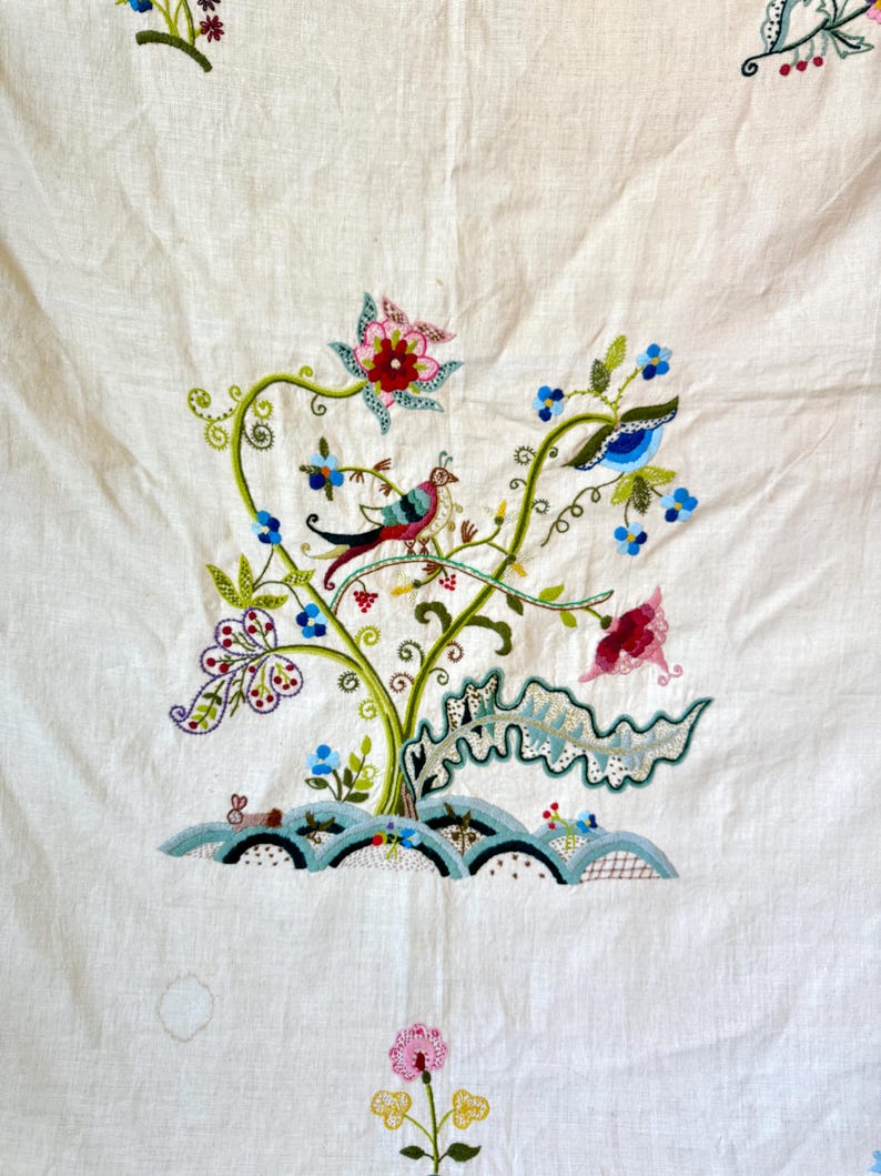May include: A white linen tablecloth with colorful embroidered flowers and birds. The embroidery features a green vine with pink, red, and blue flowers, and two birds with blue, red, and green feathers.