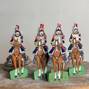 May include: Four vintage wooden toy soldiers on horseback. Each soldier is painted with a detailed uniform in blue, red, and white, mounted on a brown horse. The horses stand on green rectangular bases, creating a historical military theme.