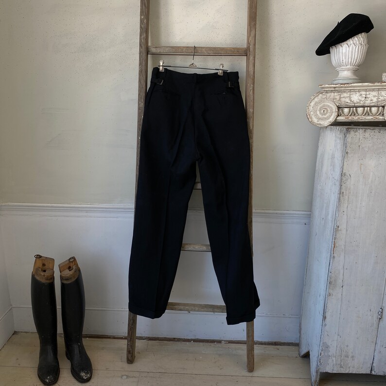 May include: A pair of dark blue, pleated trousers hanging on a wooden ladder. The trousers have a straight leg and a cuffed hem.