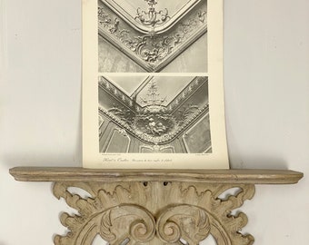 May include: A vintage architectural print titled "Les Vieux Hotels de Paris" showing detailed black and white images of ornate ceiling details. The print rests on a carved, light-coloured wooden shelf with decorative scrollwork.