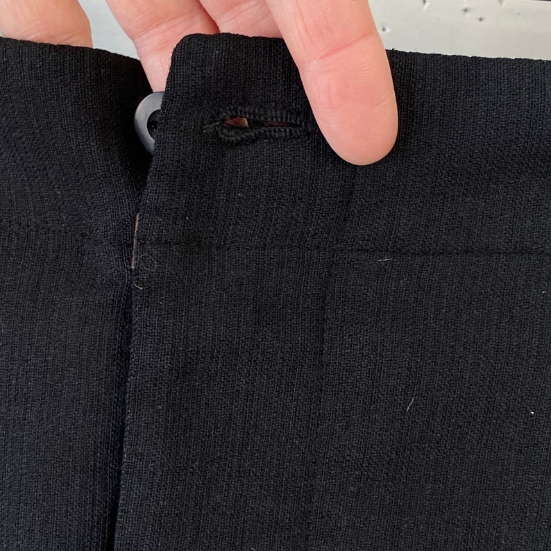 May include: Close-up of a black fabric waistband with a button and loop closure.