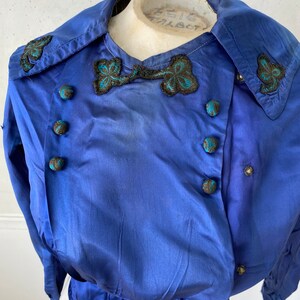 May include: A vintage cobalt blue jacket with a double-breasted design. The jacket features decorative black and teal embroidered accents on the collar and a bow-like detail. Round, teal-accented buttons adorn the front.