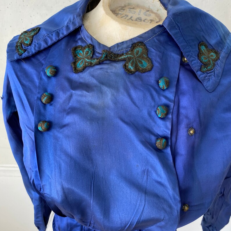 May include: A vintage cobalt blue jacket with a double-breasted design. The jacket features decorative black and teal embroidered accents on the collar and a bow-like detail. Round, teal-accented buttons adorn the front.
