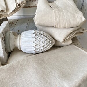 May include: A white, carved, pineapple-shaped finial sits on top of a wooden post. The finial is surrounded by several layers of beige linen fabric.