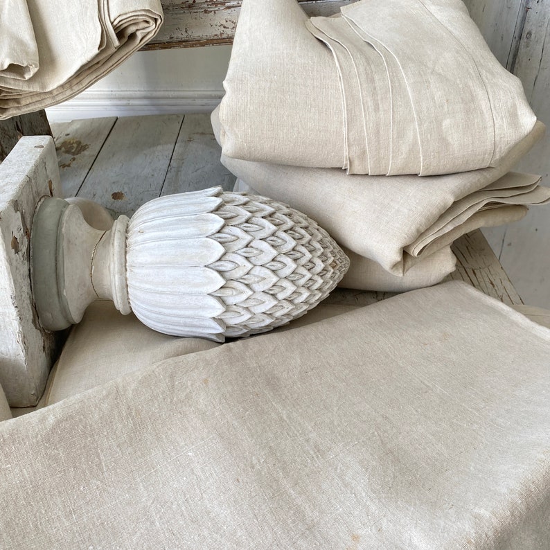 May include: A white, carved, pineapple-shaped finial sits on top of a wooden post. The finial is surrounded by several layers of beige linen fabric.