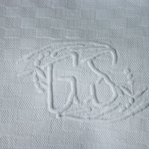 May include: White fabric with a raised embroidered design of the letters "GS" surrounded by a floral motif.