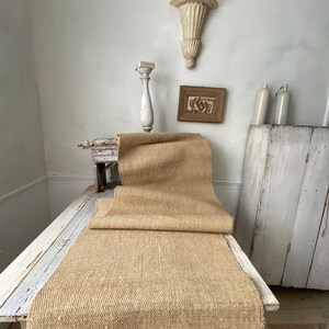May include: A large, natural, woven, beige rug is folded and displayed on a white painted table. The rug is made of natural fibers and has a rough texture. The table is distressed and has a white painted finish.