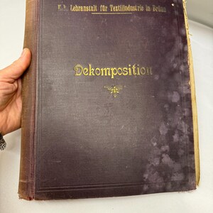 May include: A dark brown book with gold lettering on the cover. The title is "Dekomposition" and the book is from the K.k. Lehranstalt fur Textilindustrie in Brunn.