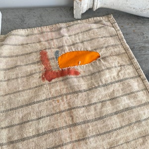 May include: A beige striped fabric with a patch of orange and red fabric sewn on. The patch has a rough, hand-stitched appearance.