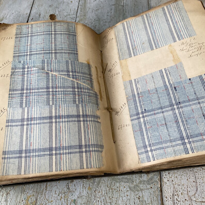 May include: An open book with fabric swatches attached to the pages. The swatches are various shades of blue and white plaid. The book has handwritten numbers and text on the pages.