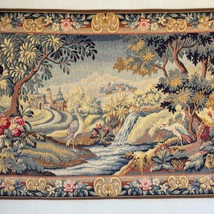 May include: A tapestry depicting a serene landscape with a waterfall, trees, and a castle. Two birds stand near a stream. The border features floral and scroll designs in shades of brown, beige, pink, and blue. The tapestry is rectangular.