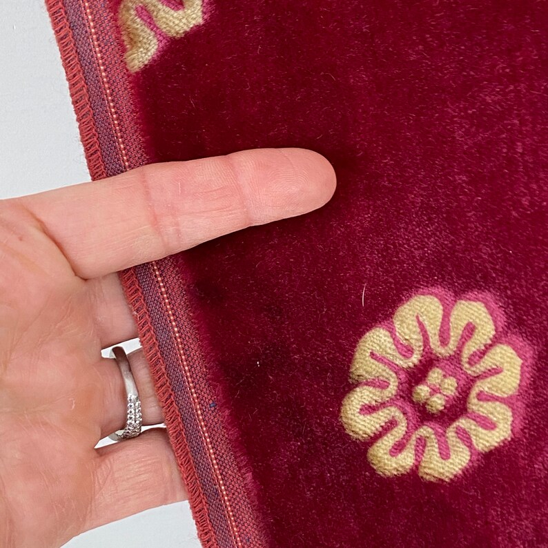以下が含まれることがあります： 金色の刺繍の花柄が施された、バーガンディ色のベルベット生地のクローズアップ。端には、バーガンディとゴールドのボーダーが付いた装飾的なトリムがあります。生地は手で持たれており、素材の質感と詳細を示しています。