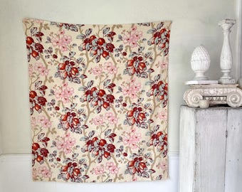 May include: A cream-colored fabric with a repeating pattern of red and pink flowers and fruit. The fabric is hanging on a white wall.