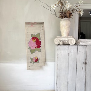 Puede incluir: Una pieza de arte textil floral vintage con una gran peonía rosa y roja con hojas verdes y un capullo más pequeño. La obra de arte se exhibe en una pared de color claro. También se ven un gabinete blanco desgastado y un jarrón con flores.