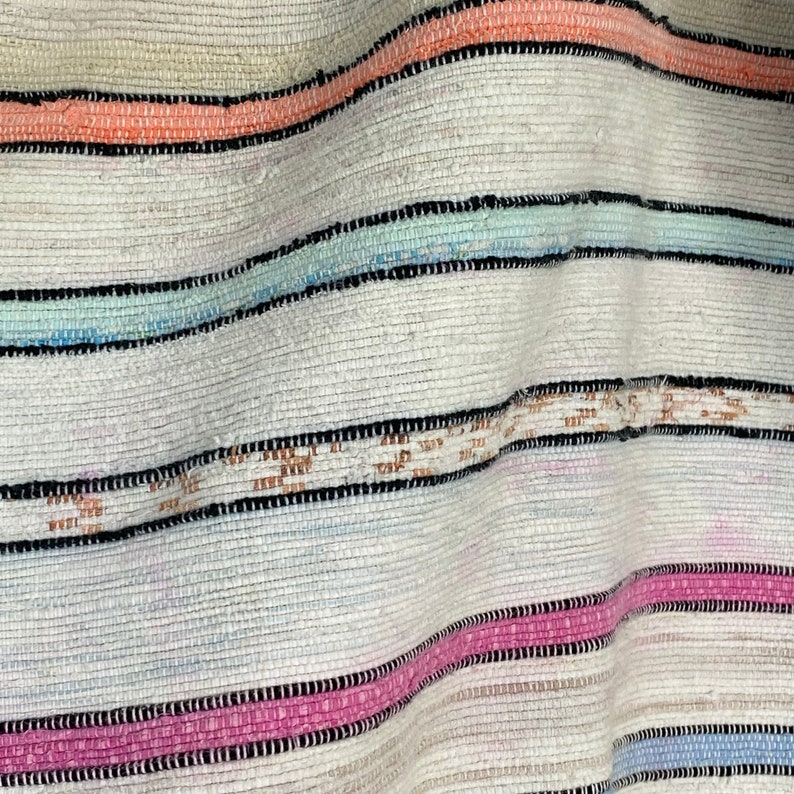 May include: A white woven rug with stripes of pink, blue, and orange. The rug is made of a soft, textured material and has a slightly distressed look.