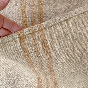 May include: Close-up of a beige linen fabric with two thick, horizontal, orange stripes. The fabric has a woven texture and a subtle diagonal pattern.