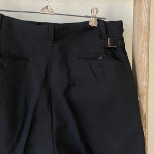 May include: Black pinstripe pants with a button and a buckle on the back. The pants have two pockets on the back.