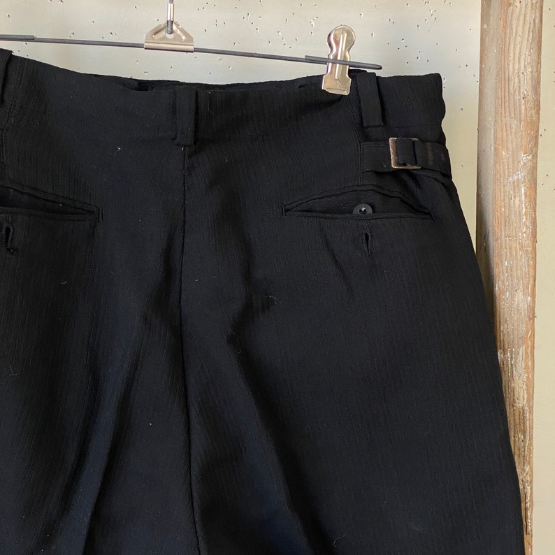 May include: Black pinstripe pants with a button and a buckle on the back. The pants have two pockets on the back.