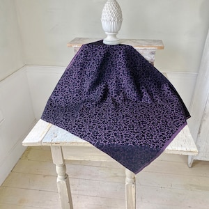 May include: A purple and black patterned fabric with a small, repeating design. The fabric is draped over a white table.