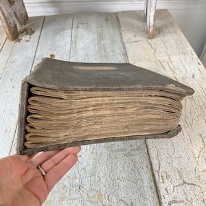 May include: A vintage, dark gray, bound book with a worn leather cover and aged, yellowed pages. The book is closed and lying on a white, weathered wooden surface.