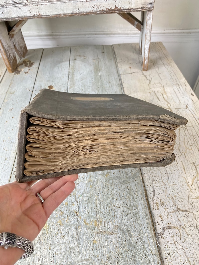 May include: A vintage, dark gray, bound book with a worn leather cover and aged, yellowed pages. The book is closed and lying on a white, weathered wooden surface.