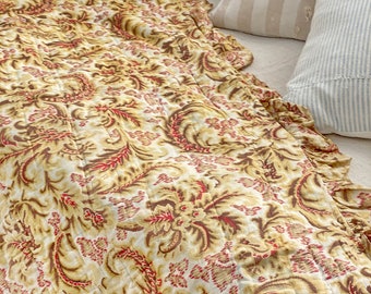 May include: A cream-colored quilted throw blanket with a red and brown floral pattern. The blanket has a ruffled edge and is made of a soft, lightweight fabric.