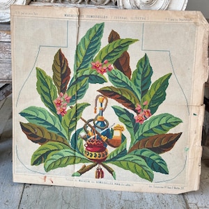 May include: A vintage needlepoint pattern featuring a floral design with green leaves, pink flowers, and a blue and red smoking pipe with a red and gold base. The pattern is printed on a white background with the text "MAGASINDES DEMOISELLES (JOURNAL ILLUSTRE)" at the top.