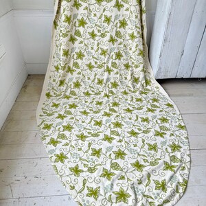 May include: A white oval tablecloth with a green and white grapevine pattern. The tablecloth is made of a soft, natural fabric and has a subtle sheen.