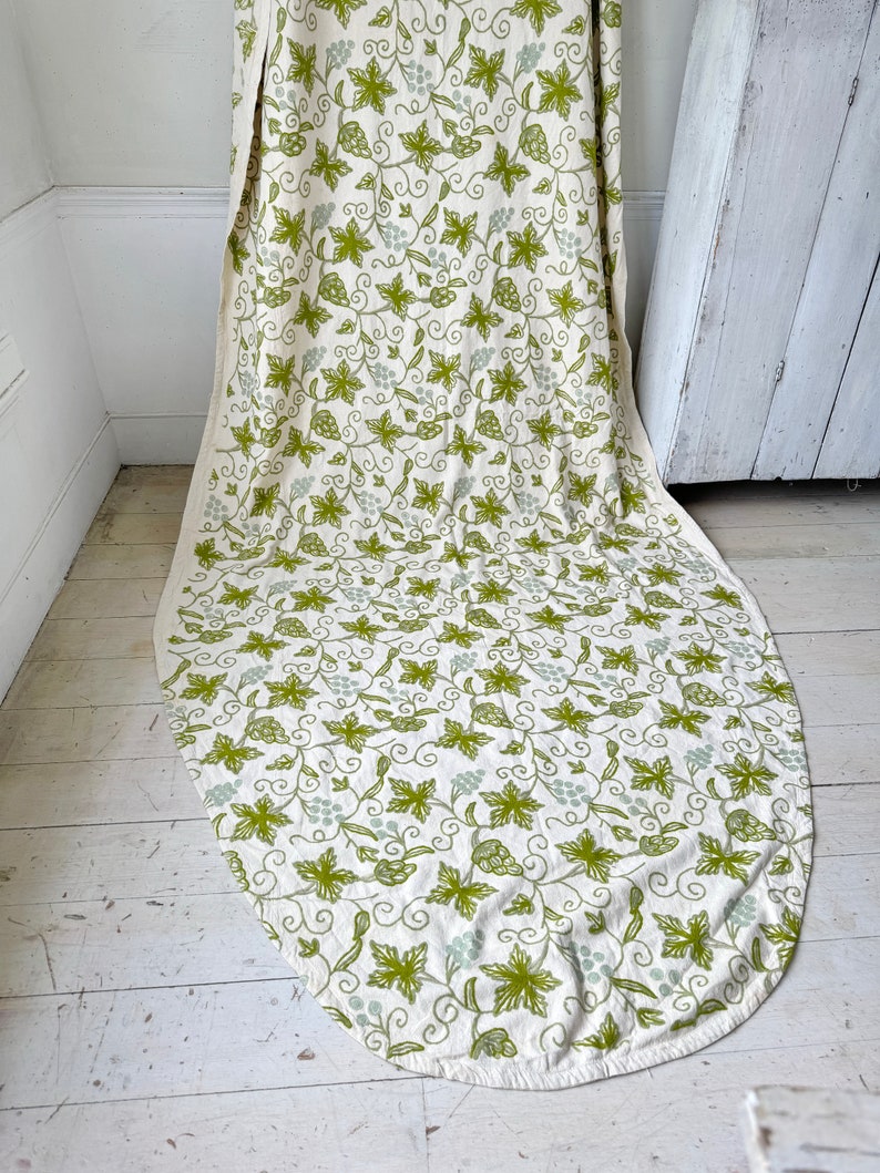 May include: A white oval tablecloth with a green and white grapevine pattern. The tablecloth is made of a soft, natural fabric and has a subtle sheen.