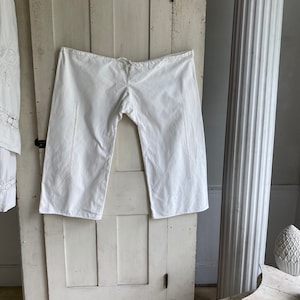 May include: White cotton pants with embroidered details hanging on a white wooden door. The pants have a drawstring waist and are cropped.