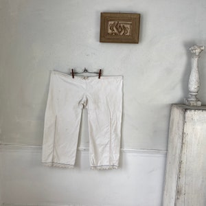 Puede incluir: Pantalón blanco de algodón con ribete de encaje en la parte inferior. El pantalón está colgado de una cuerda para tender la ropa.