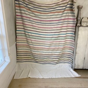 May include: A white woven tapestry with colorful stripes in shades of pink, blue, green, and brown. The tapestry has a white fringe at the bottom.