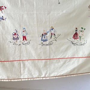May include: A white tablecloth with red stitching around the edges. The tablecloth is embroidered with colorful figures in traditional French clothing. The text "Ile de France", "Bourdonnais", and "Franche Comte" is embroidered on the tablecloth.