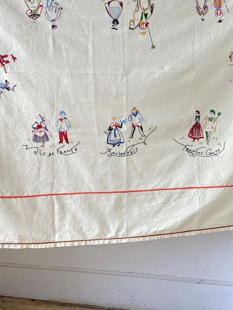 May include: A white tablecloth with red stitching around the edges. The tablecloth is embroidered with colorful figures in traditional French clothing. The text "Ile de France", "Bourdonnais", and "Franche Comte" is embroidered on the tablecloth.