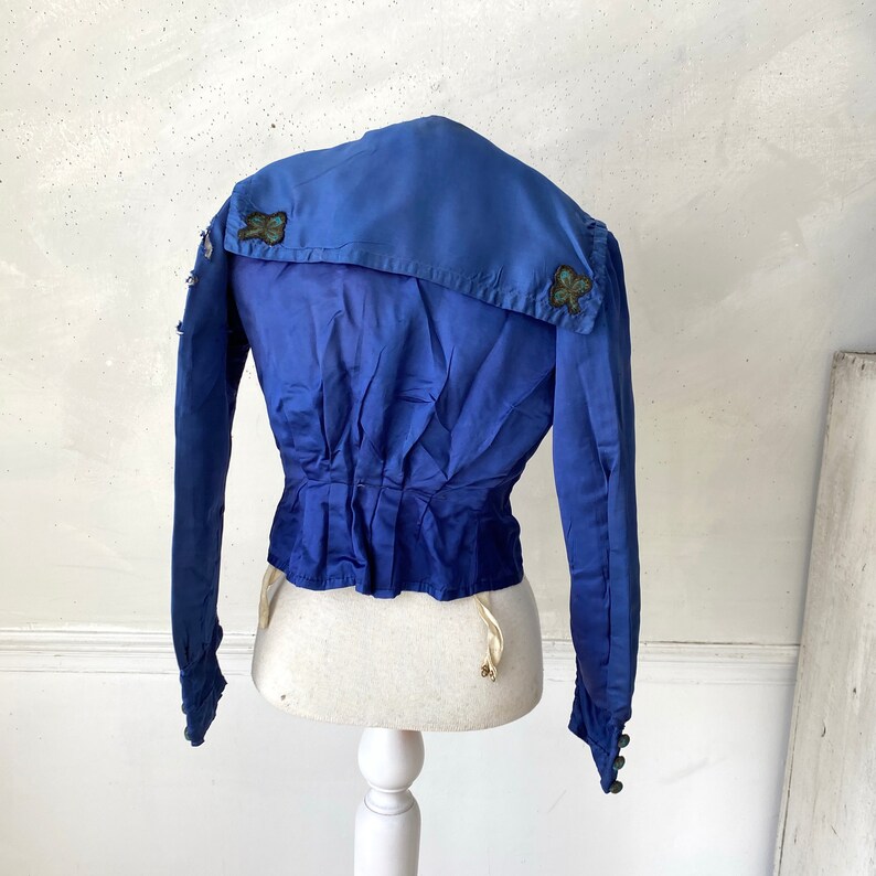 May include: A vintage cobalt blue jacket with long sleeves and a sailor collar. The back of the jacket is gathered at the waist and features decorative embellishments on the collar. The jacket is displayed on a white mannequin.