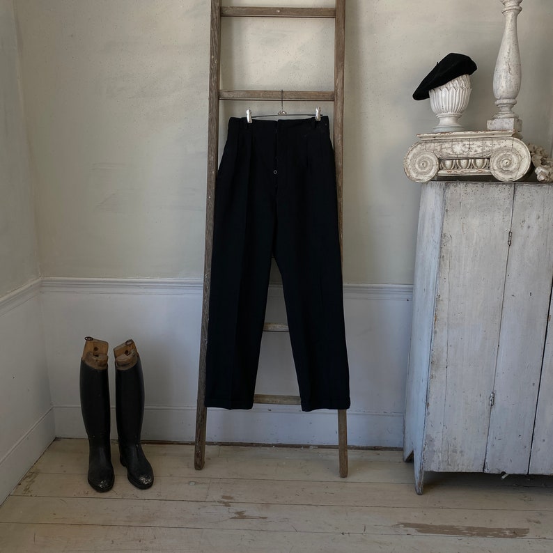 May include: Black high-waisted pants with pleats hanging on a wooden ladder. Two black leather boots are on the floor below.