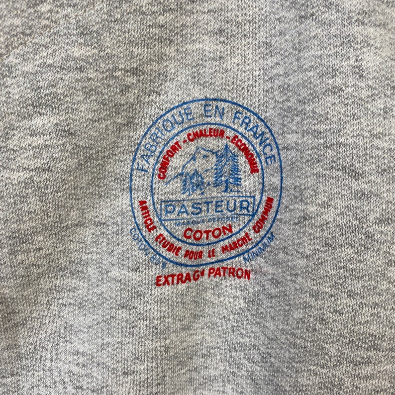 Pode incluir: Uma camisola cinzenta com um logotipo circular azul e vermelho que diz "Fabrique en France" e "Pasteur" com uma cena de montanha e o texto "Confort - Chaleur - Economie" e "Article Etudie Pour le Marche Commun" e "Coton 85$" e "Extra-G Patron".
