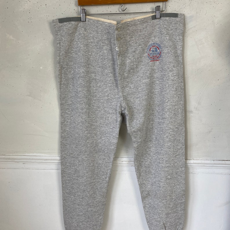 Pode incluir: Cal&ccedil;as de moletom cinzas com c&oacute;s branco e fechamento com bot&atilde;o. As cal&ccedil;as de moletom t&ecirc;m um logotipo circular na perna direita que diz "Champion" e "Athletic Wear".