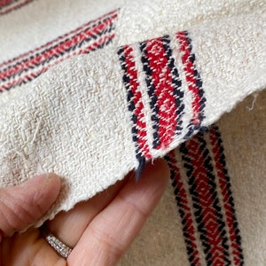 May include: Close-up of a white linen fabric with red and black woven stripes. The fabric has a rough texture and is folded over.