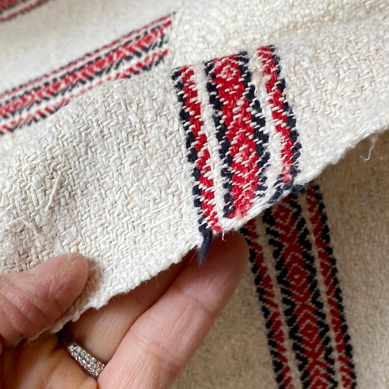 May include: Close-up of a white linen fabric with red and black woven stripes. The fabric has a rough texture and is folded over.