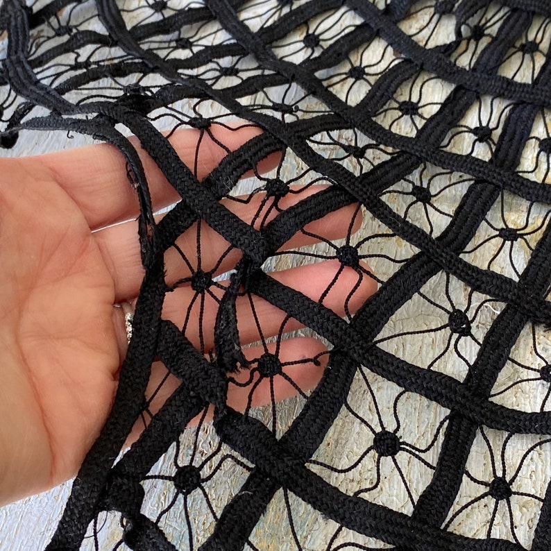 May include: Black lace fabric with a geometric pattern of squares and circles. The fabric is made of a thick, woven material and has a slightly textured surface.