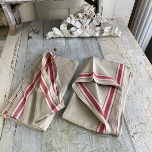 Pode incluir: Duas toalhas de chá de linho com listras vermelhas. As toalhas estão dobradas e colocadas sobre uma superfície de madeira branca.