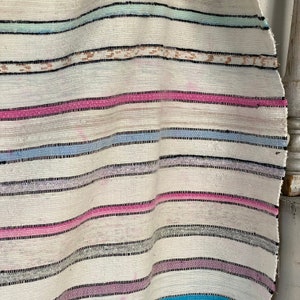 May include: A white woven rug with pink, blue, and gray stripes. The rug is made of a soft, natural material and has a slightly distressed look.