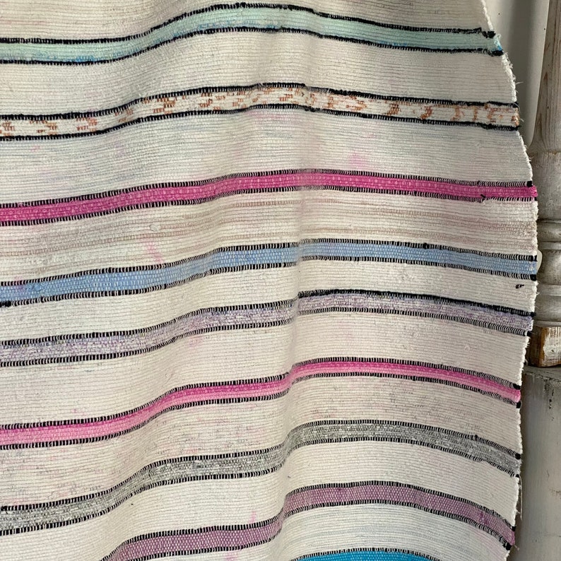 May include: A white woven rug with pink, blue, and gray stripes. The rug is made of a soft, natural material and has a slightly distressed look.