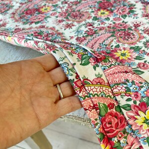 May include: Close-up of a floral fabric with a white background. The fabric features a paisley design with pink, red, yellow, and blue flowers. The fabric is folded, revealing the pattern and texture.