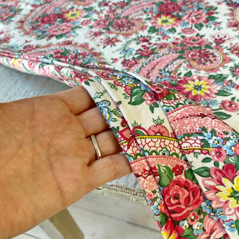 May include: Close-up of a floral fabric with a white background. The fabric features a paisley design with pink, red, yellow, and blue flowers. The fabric is folded, revealing the pattern and texture.