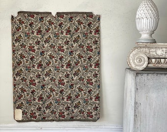 May include: A vintage floral tapestry with a white background and a repeating pattern of red, pink, and blue flowers. The tapestry is framed by a wooden border and is hanging on a white wall.
