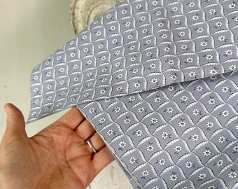 May include: A section of fabric with a repeating floral pattern in light blue and white. The fabric is held in a hand, revealing the detailed design. The pattern features small white flowers within a grid-like structure, creating a vintage aesthetic.
