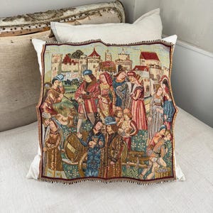 May include: A decorative pillow with a medieval tapestry design. The tapestry depicts a scene with figures in period clothing, a castle, and a cart. The pillow has a white background and a fringed border.
